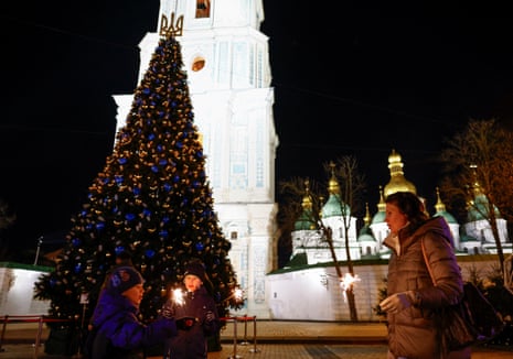 یک زن با کودکان در جشن سال نو در مقابل کلیسای جامع سنت سوفیا در کیف، جرقه‌هایی را در کنار درخت کریسمس نگه می‌دارد.